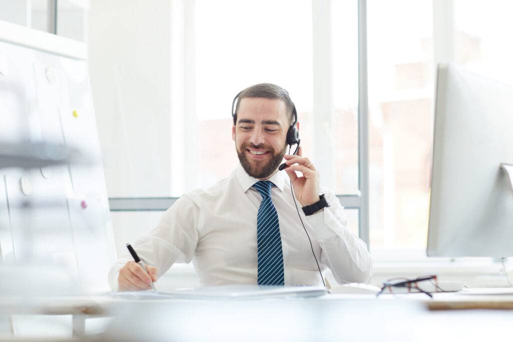 Positive communicative handsome young call center agent sitting at table and making notes in notepad while talking to customer via headset